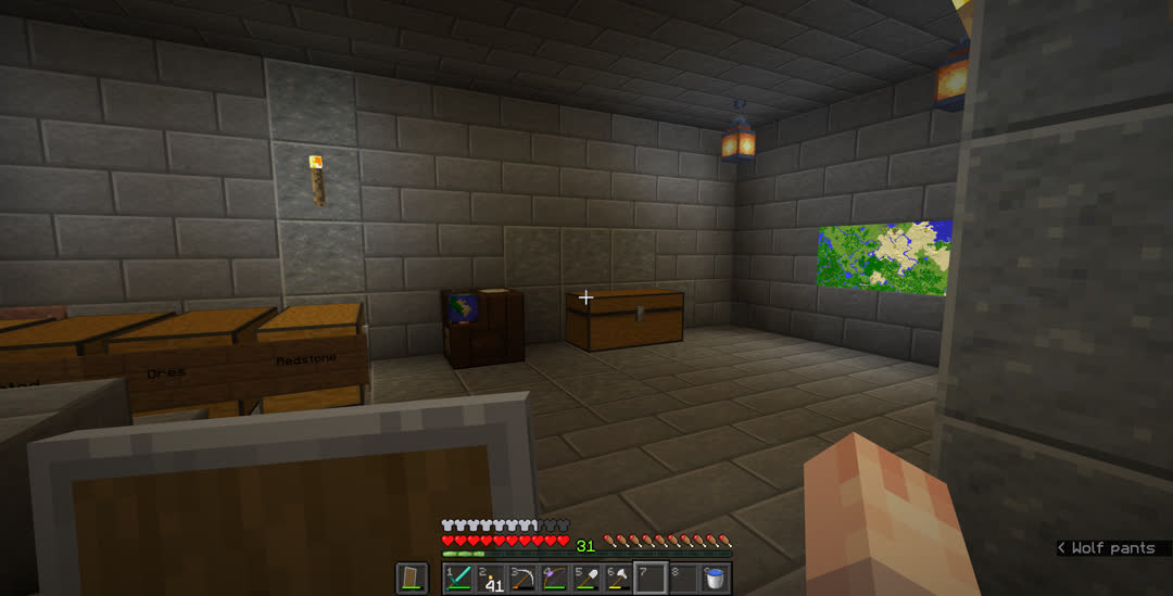 The subbasement now has stone bricks for its walls and floor, except for a few columns of polished andesite and patches of that and polished granite. A few chests are against the wall at the bottom of the stairs, with the labels “Ores” and “Redstone” visible. Along with a cartography table and unlabeled chest, there are two backlit maps on the far wall.